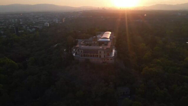 Aerial panning view Mexico City Castle with tower in middle surrounded by Chapultepec forest and cityscape with sunset over horizon background
