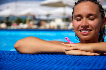 Calm happy girl with her eyes closed enjoys her vacation, being in the warm water of the pool at the resort. Portrait of a beautiful young woman with a smile in the pool on vacation