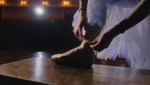 Ballet female dancer puts on pointe shoes preparing for choreography rehearsal. Ballerina practices before performance and dances on tiptoe on theater stage illuminated by spotlights. Ballet show.