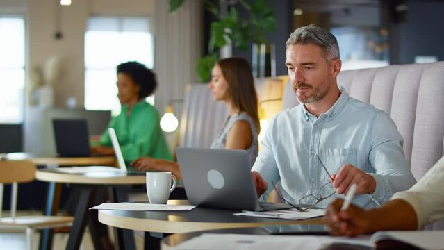 Mature businessman wearing glasses working on laptop in informal seating area of modern office on coffee break - shot in slow motion