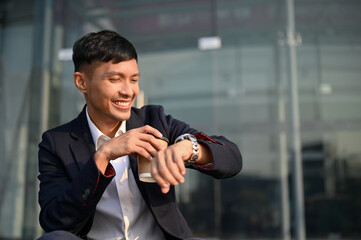 Happy Asian businessman checks the time, looking at his watch, while sipping coffee