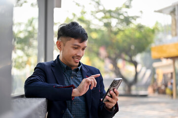 Happy Asian businessman using his smartphone while waiting for a bus or taxi in the city.