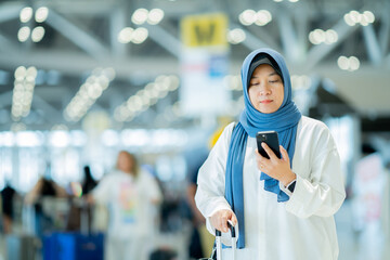 An Asian Muslim wearing a blue hijab is preparing for a vacation and she is at the airport. She is using her mobile phone to contact her friends and Muslim travelers.