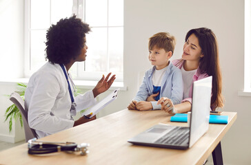 Little boy patient with his mother talking with friendly african american pediatrician doctor on consultation in her office in clinic and listening the recommendations after medical examination. - Powered by Adobe