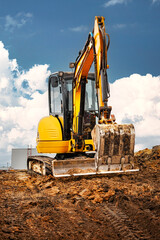 Mini excavator at the construction site on the edge of a pit against a cloudy blue sky. Compact construction equipment for earthworks. An indispensable assistant for earthworks. - obrazy, fototapety, plakaty