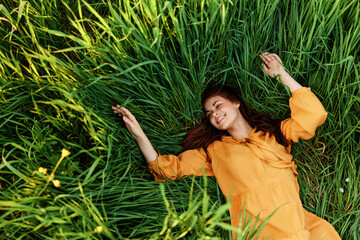 a relaxed red-haired woman enjoys summer lying in the tall green grass in a long orange dress smiling happily with her eyes closed