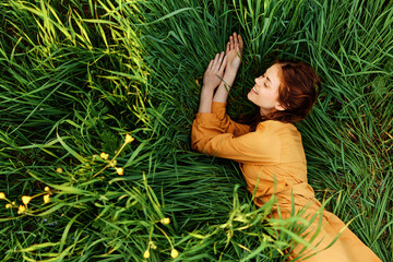 a relaxed red-haired woman enjoys summer lying in the tall green grass in a long orange dress smiling happily looking away