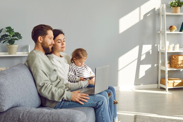Young family sitting on sofa and using laptop. Couple watching video on notebook computer while their little child is playing on modern tablet or mobile phone. Technology, modern lifestyle concept