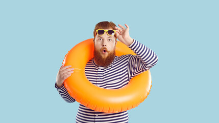 Astonished funky crazy man with inflatable swimming circle isolated on pastel turquoise background. Chubby red-bearded man in striped sailor clothes looks at camera in shock, raising his sunglasses. - obrazy, fototapety, plakaty
