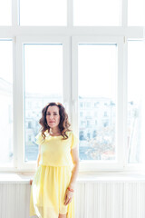 beautiful brunette woman in a yellow dress stands at a large window