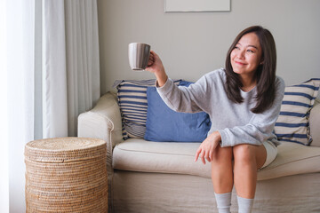 Fototapeta premium Portrait image of a young woman raising hand and say hi to friends outside while drinking coffee at home