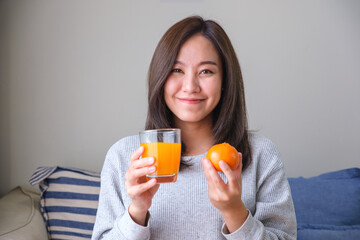 Portrait image of a young woman holding an orange and a glass of fresh orange juice at home
