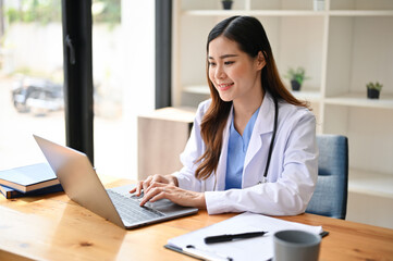 Beautiful Asian female doctor in uniform using her laptop at her workspace in a hospital office.