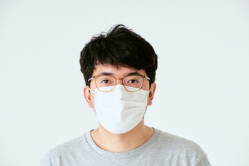 白背景でカメラ目線のマスクと眼鏡を着けた30代の1人の日本人男性の人物の上半身の正面