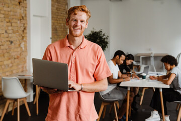 Smiling man working on laptop computer while standing in office with his colleagues on a background
