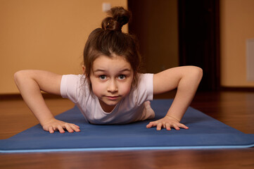 Close-up front view of Caucasian determined active little girl in white t-shirt and sportswear, looking confidently at camera, doing push ups on fitness mat at home. Child practicing yoga, exercising