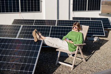 Young woman sits relaxed on a lounge chair while resting on rooftop with a solar station. Happy...