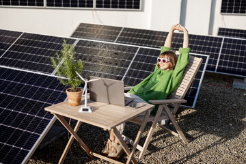 Woman works on laptop while sitting relaxed by the table on rooftop with a solar power plant. Concept of remote work, alternative energy and sustainable lifestyle