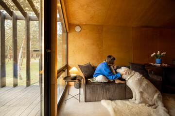 Woman cares her dog while sitting on a couch by the window, spending leisure time together at...