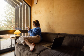Woman sitting relaxed with a phone on a couch by the window at wooden cottage in forest. Concept of...