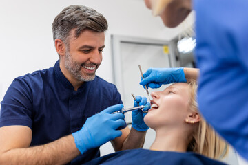 Fototapeta premium Close up of dentist's hands holding tool and syringe in hands. Pain killer in action. Female client keep mouth opened. Beautiful smile and white teeth. Cut view.