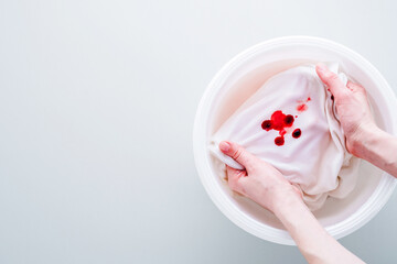 Female hands soak blood stain on the white clothes in a plastic basin of cold water. Removal of excess blood. isolated. top view. daily life stain concept