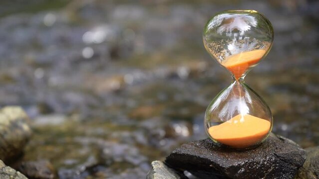 Time flows away like the water in the river. An hourglass with blur water in background. concept of time and flowing water. An hourglass on a rock of a creek.