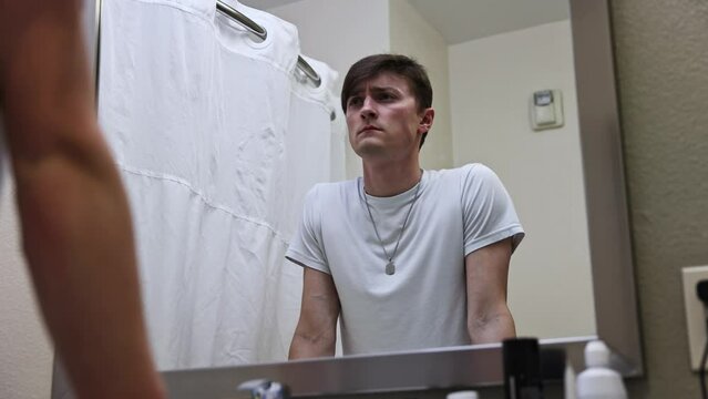 young white male in a white tee walking up to a hotel room bathroom mirror depressed and sighing in grief in the mirror reflection