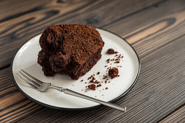 A piece of chocolate bundt cake and a dessert fork on white plate on wooden table, copy space