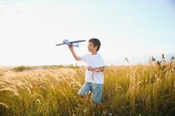 Naklejka na ściany i meble Cute happy cheerful child running fastly along grassy hill at countryside holding big toy plane in hand. Boy playing during sunset time in evening. Horizontal color photography.