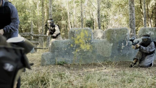 Group of men and women in protective clothes and helmets shooting with paintball markers. Teams fighting on battlefield outdoors.