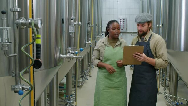 Medium wide shot of two multiethnic male and female brewery employees standing in fermentation room among large steel bright beer tanks, looking at gauges and completing checklist on clipboard