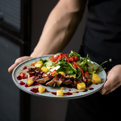 Meat steak cut into pieces with fresh vegetable salad, cranberry sauce and pineapple. Dish on blue plate in hands of waiter. Lunch at restaurant. View from above. 