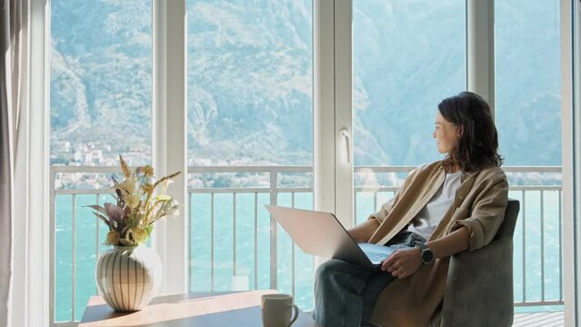 A young caucasian woman relaxing and enjoying a beautiful view of the bay and mountains while sitting in a living room and working on a laptop.