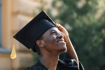 Portrait closeup of afro american student in graduation mantle and hat standing outdoors and with pleasure looking up. Job search, graduate from university, start in life, thoughts about future. - obrazy, fototapety, plakaty