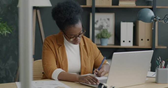 Portrait of business owner wearing glasses using laptop typing and writing financial imformation in notebook working in office. Job and workplace concept.