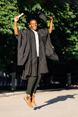 Joyful african american graduate from university with big smile jumping up in black mantle and hat....