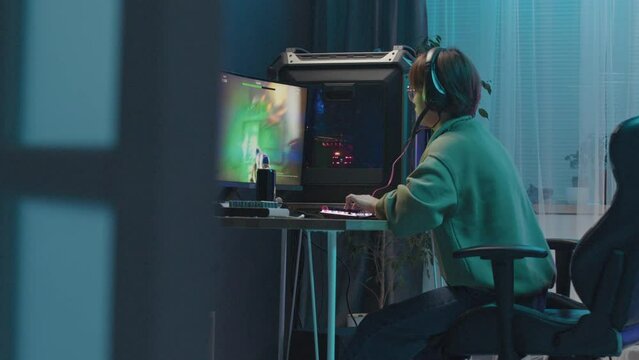 Full side shot of unrecognisable gamer sitting at desk late at night and playing FPS online videogame on computer, firing bullets from gun and hitting targets