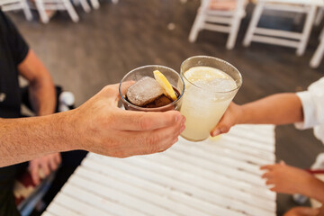 Two hands holding glasses with refreshing drinks toasting in restaurant at night
