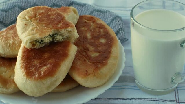A glass of fresh milk and a plate with ruddy, golden, fried homemade pies, one pie is broken in half and one can see the filling of cottage cheese with herbs. Close-up. The concept of home baking