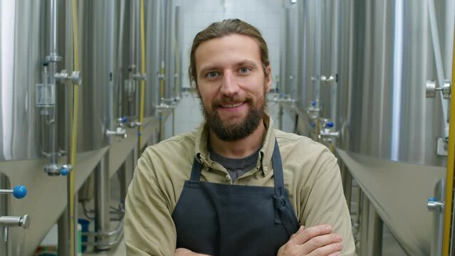 Medium close-up portrait shot of young male Caucasian craft brewery owner with red hair and beard, in apron standing between beer fermentation tanks and looking at camera with confident smile