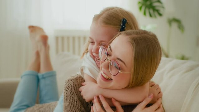 Caucasian little daughter embrace mother by neck piggyback cuddling hugging babysitter woman mom with preschool child kid baby smiling laughing lying on cozy couch at home happy family at home bonding