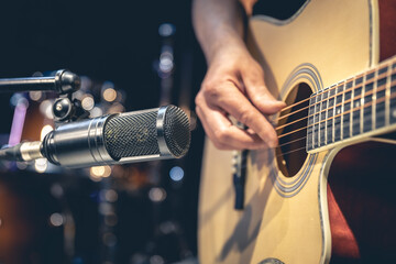 Male musician playing acoustic guitar behind microphone in recording studio. - obrazy, fototapety, plakaty
