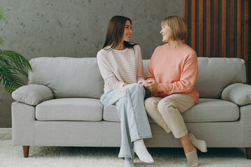 Full body two adult women mature mom young kid wear casual clothes look camera talk speak hug sit on gray sofa couch stay at home flat rest relax spend free spare time in living room. Family concept.