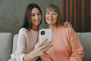 Two adult happy fun women mature mom young kid wear casual clothes hold use mobile cell phone sit on gray sofa couch stay at home flat rest relax spend free spare time in living room. Family concept.