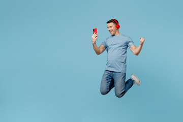Full body young smiling happy fun man wear casual t-shirt jump high use mobile phone do winner gesture listen music in headphones isolated on plain pastel light blue cyan background studio portrait.