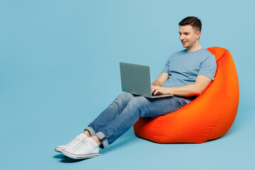 Full body young smiling happy IT man wears casual t-shirt sit in bag chair hold use work on laptop pc computer isolated on plain pastel light blue cyan background studio portrait. Lifestyle concept