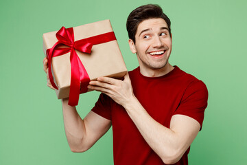 Young smiling happy fun cheerful man he wearing red t-shirt casual clothes holding present box with gift ribbon bow isolated on plain pastel light green background studio portrait. Lifestyle concept.