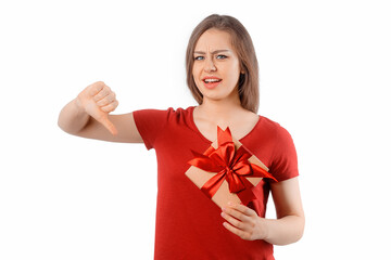Fototapeta premium Portrait of a winsome girl holding gift box, showing thumb down. Unhappy brunette woman with long hair and frowning with disappointed face, stand over white background.