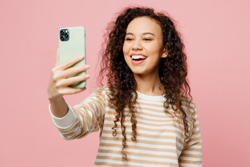 Young woman of African American ethnicity she wears light casual clothes doing selfie shot on mobile cell phone post photo on social network isolated on plain pastel pink background studio portrait.